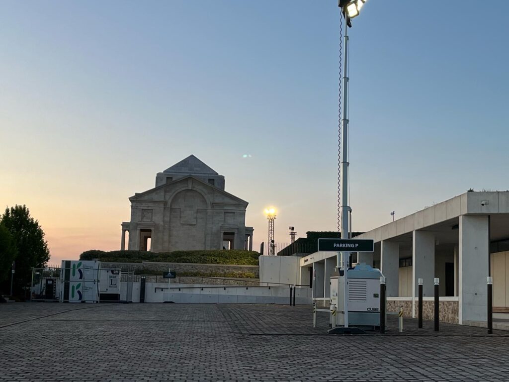 Mât d'éclairage mobile CUBE LED Revolt Location déployé sur le parking d'un site patrimonial, à proximité de panneaux Revolt Energygreen, au coucher du soleil.