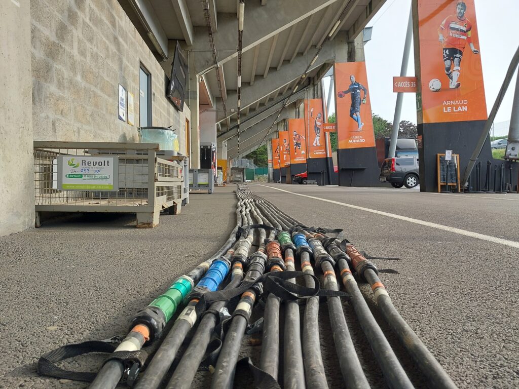 Faisceau de câbles électriques industriels multicolores déployés sous les tribunes du stade de Lorient avec caisse de matériel Revolt Location visible à gauche