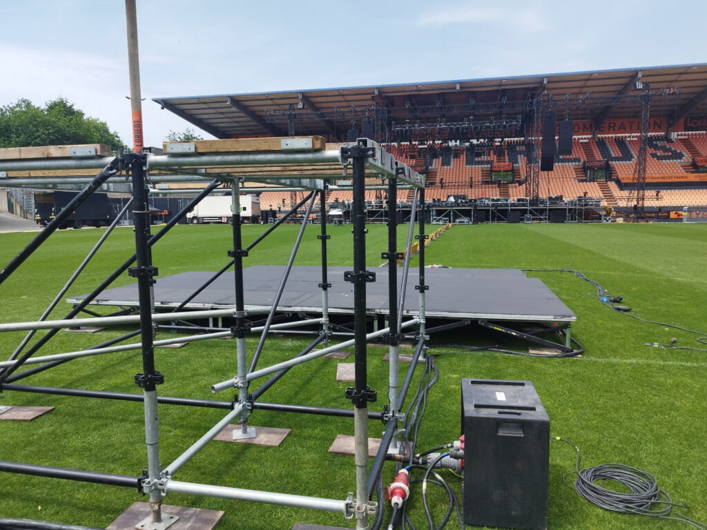 Installation d'une scène événementielle sur la pelouse du stade de Lorient avec câblage électrique et coffret de distribution au premier plan – Revolt Location