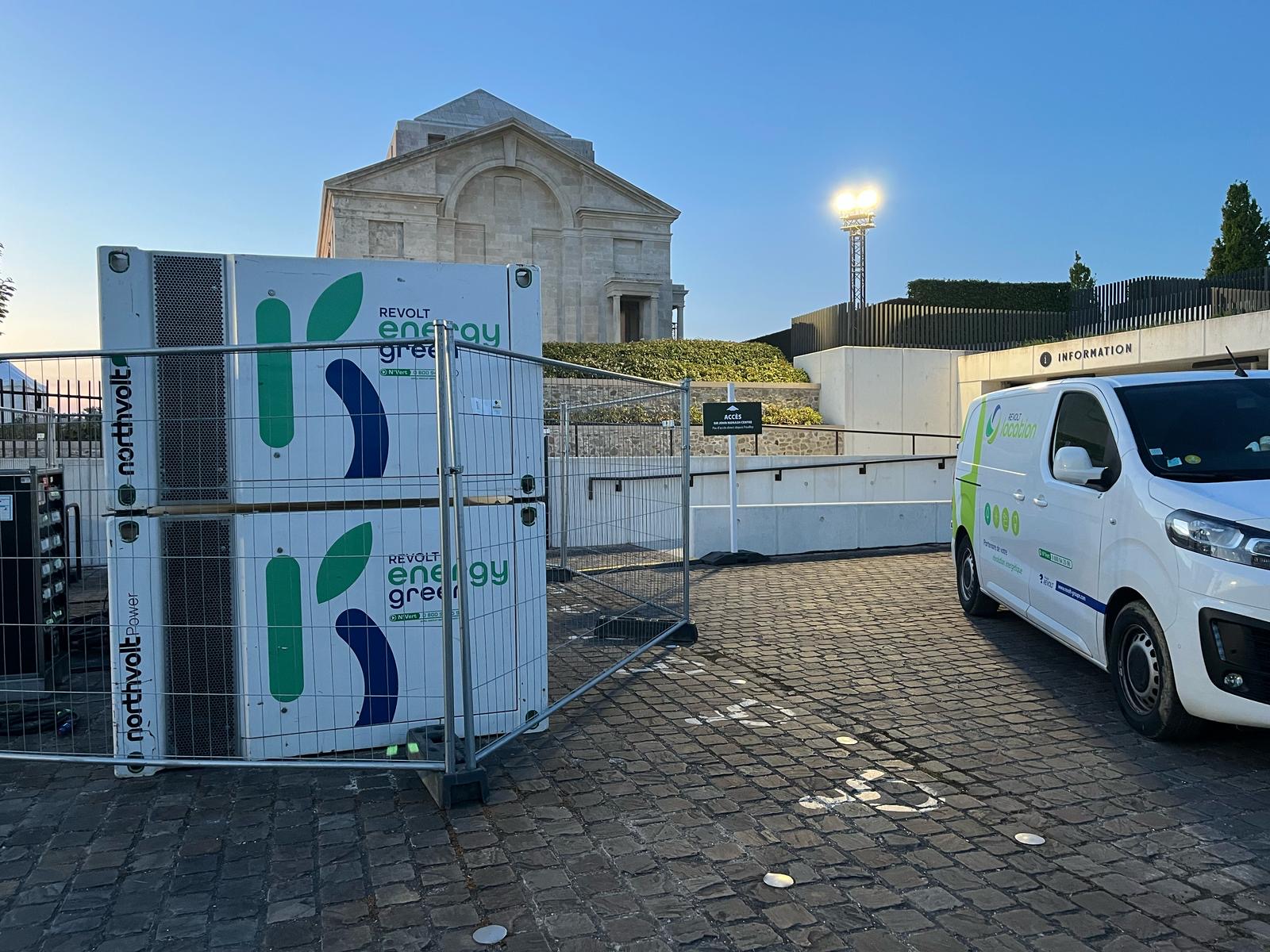 Packs batteries Revolt Energygreen Northvolt sécurisés par une clôture de chantier, avec véhicule utilitaire Revolt Location stationné devant un site patrimonial.