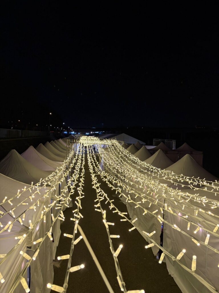 Guirlandes lumineuses décoratives Revolt Location suspendues au-dessus d'une allée bordée de chapiteaux blancs, illuminant un site événementiel de nuit.
