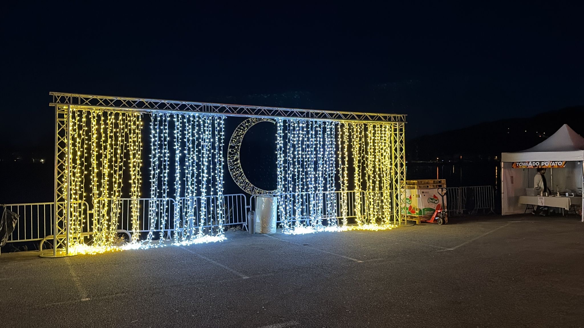 Rideau lumineux décoratif Revolt Location bicolore monté sur structure aluminium, orné d'un motif lumineux en forme de lune, installé sur un site événementiel nocturne en bord d'eau.