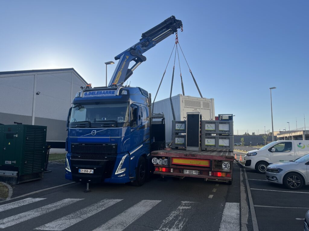 Camion-grue Volvo FH déchargeant un container de stockage d'énergie sur plateau – livraison et installation de matériel électrique par Revolt Location