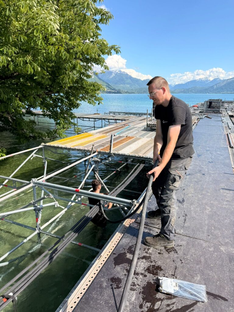 Technicien Revolt Location tirant un câble électrique sur une scène flottante au bord du lac d'Annecy lors du montage du Festival International du Film d'Animation – installation en milieu aquatique