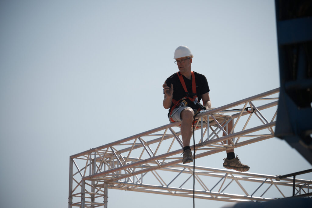 Technicien Revolt Location casqué et harnaché, assis en hauteur sur une structure aluminium lors d'une opération de montage technique.