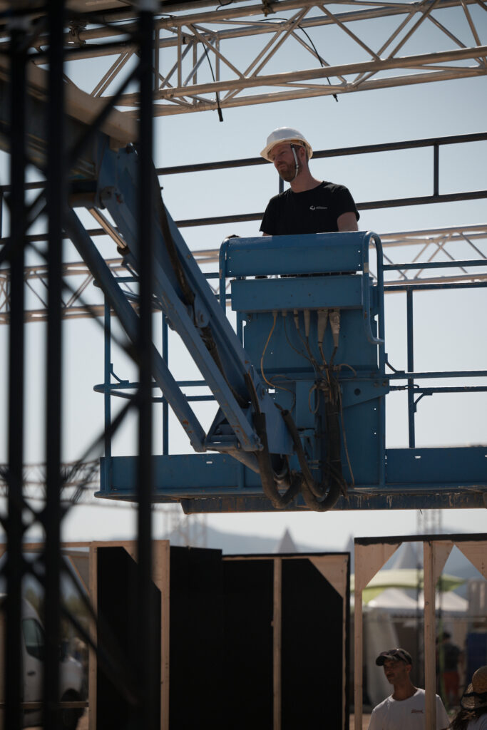 Technicien Revolt Location casqué, positionné dans une nacelle élévatrice, intervenant sous une structure aluminium en hauteur sur un site événementiel.