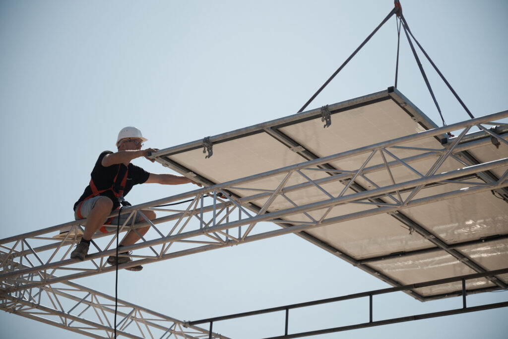 Technicien casqué et harnaché guidant à la main la pose d'un module photovoltaïque suspendu à une grue sur une structure aluminium en hauteur.