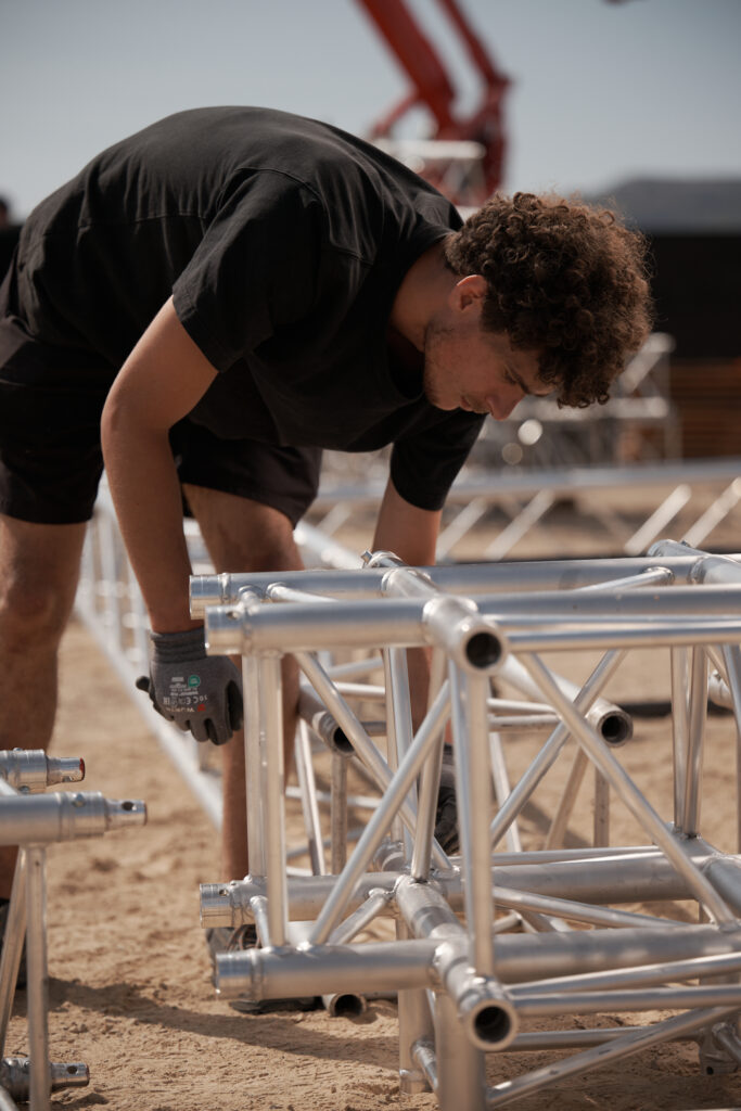 Technicien Revolt Location ganté assemblant des éléments de structure en treillis aluminium au sol, sur un chantier événementiel.