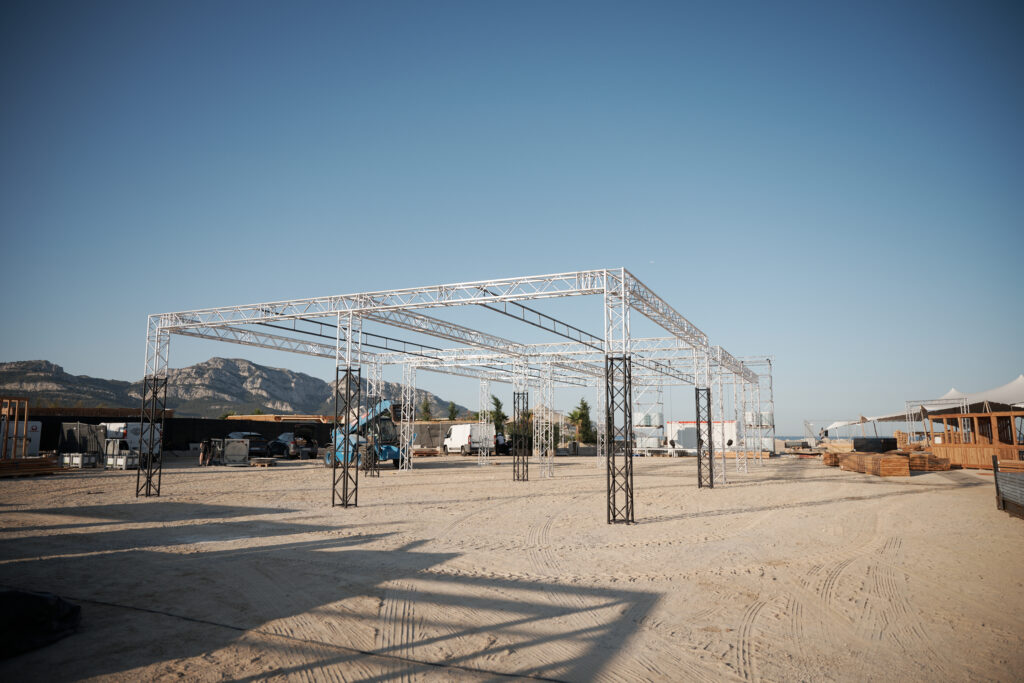 Structure en trusses aluminium en cours de montage sur une plage en bord de mer, support technique pour un événement extérieur.