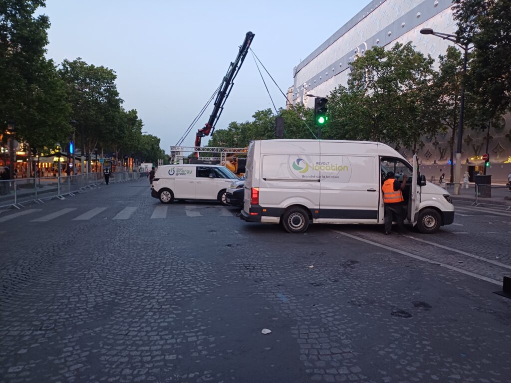 Utilitaires Revolt Location et Revolt Energy Green stationnés sur les Champs-Élysées près d'une grue de montage, technicien intervenant sur site.