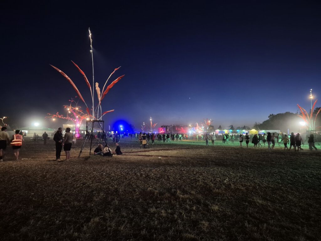 Site de festival au crépuscule avec structures lumineuses décoratives, stands illuminés et public traversant l'espace – éclairage événementiel Revolt Location.