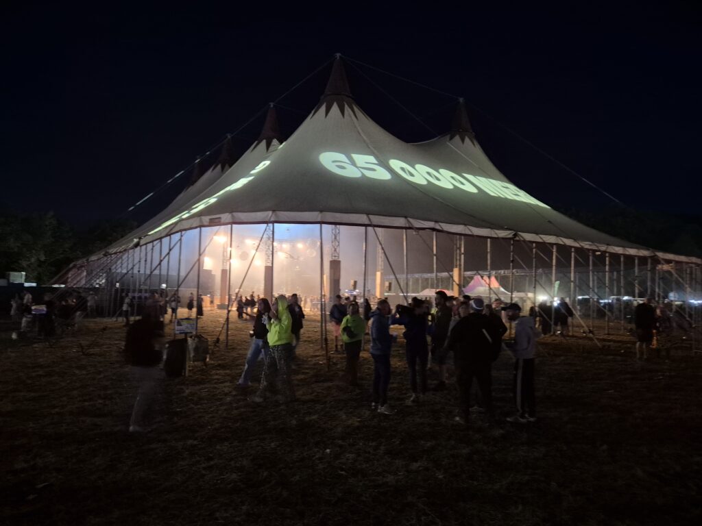 Chapiteau de concert nocturne avec projection numérique « 65 000 » sur la toile et public rassemblé devant la scène éclairée – installation événementielle Revolt Location.