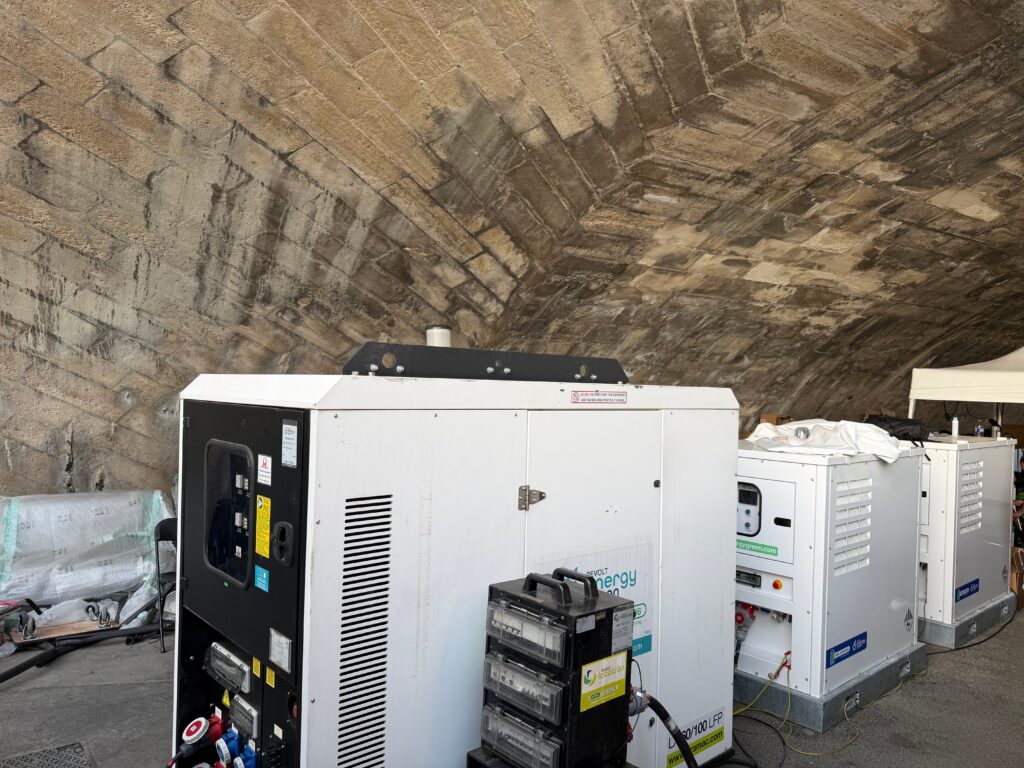 Unités de stockage d'énergie Revolt Energy Green alignées sous une voûte en pierre, avec coffret de comptage électrique au premier plan.