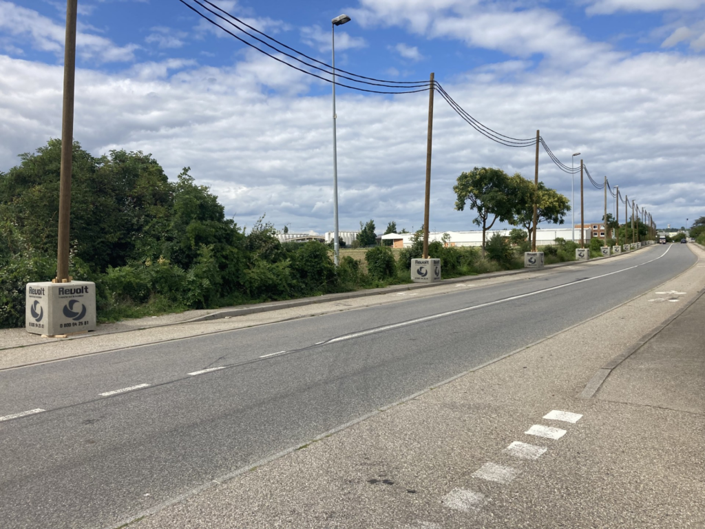 Poteaux d'une ligne aérienne électrique Revolt Location, ancrés par des plots béton floqués, alignés le long d'une route en zone industrielle.