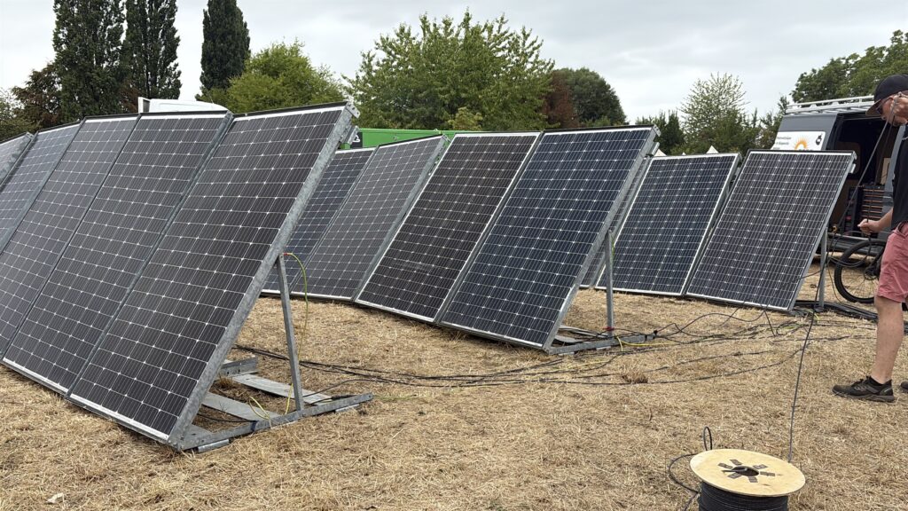 Rangées de panneaux photovoltaïques sur supports galvanisés inclinés, câbles au sol et intervenant déroulant un câble depuis un touret, sur un terrain sec.