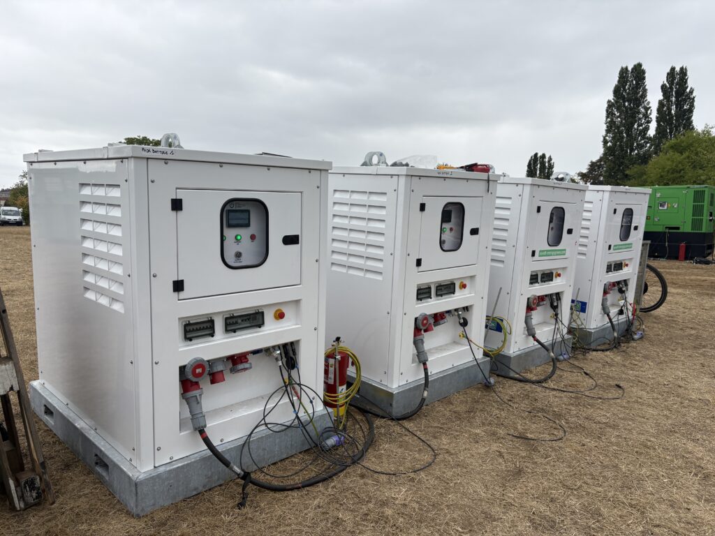 Alignement de quatre packs batteries mobiles blancs avec écran de contrôle, arrêt d'urgence et prises industrielles rouges, installés sur un terrain herbeux.