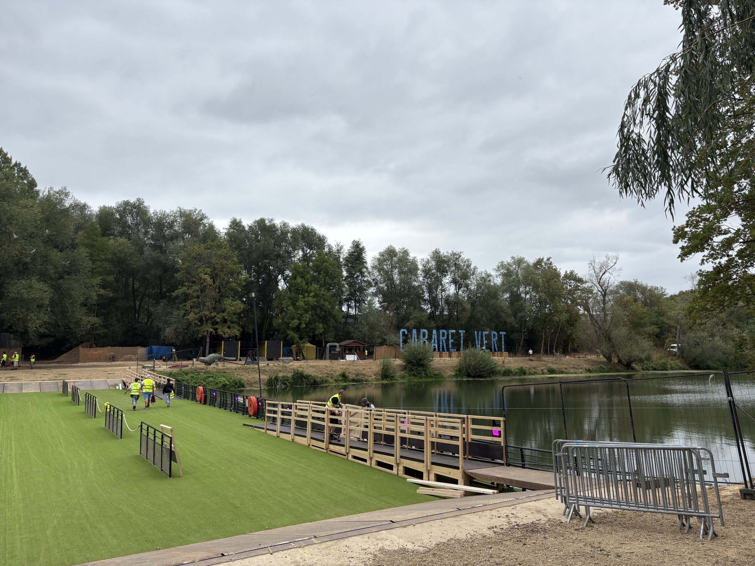 Montage du site du festival Cabaret Vert : passerelle en bois au bord d'un plan d'eau, pelouse synthétique et équipes en gilets jaunes – Revolt Location.