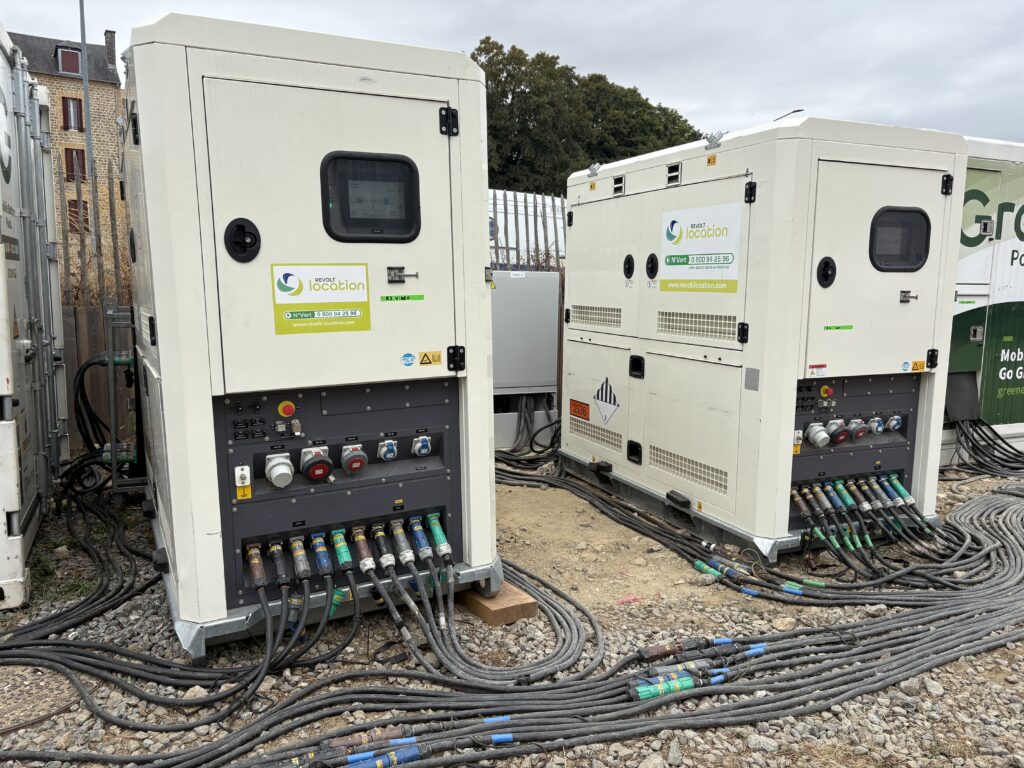 Deux unités de stockage d'énergie blanches Revolt Location avec écran de contrôle, prises industrielles et câbles raccordés, sur terrain gravillonné.