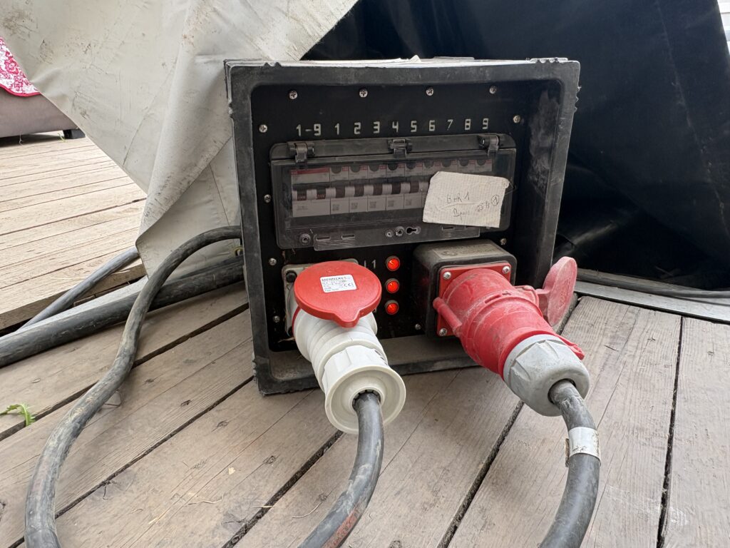 Armoire de distribution électrique noire avec disjoncteurs numérotés et deux prises industrielles raccordées, sur un plancher en bois – Revolt Location.