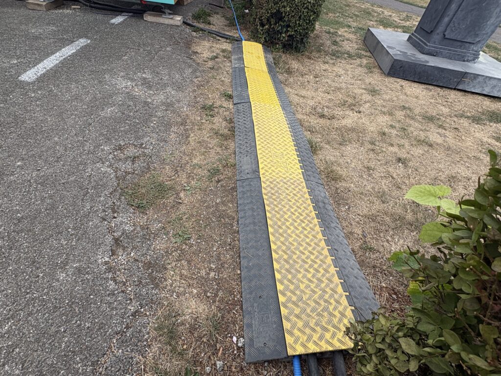 Passage de câble jaune et noir posé sur une pelouse, protégeant des câbles électriques noirs et bleus à côté d'une voirie – Revolt Location.